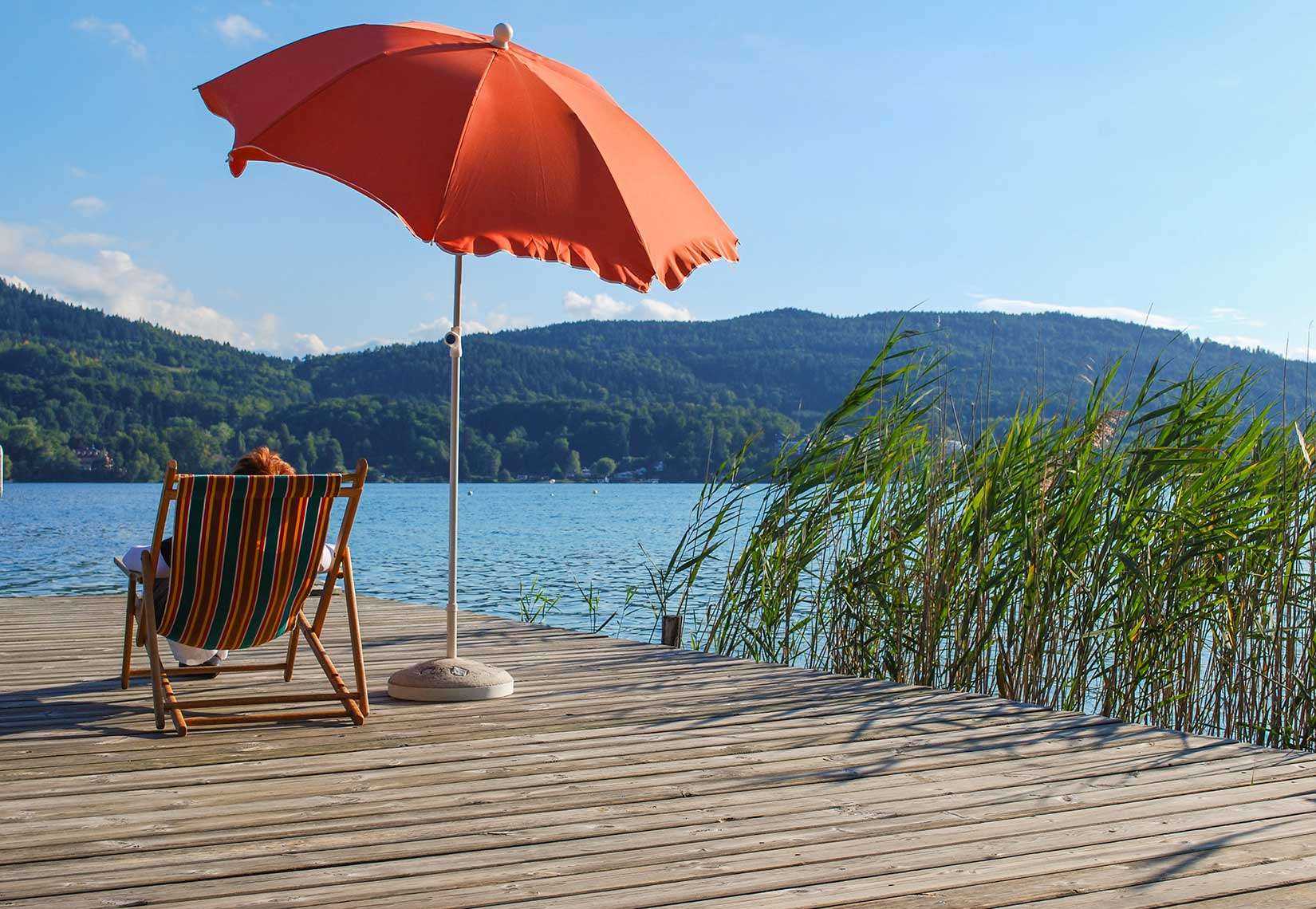 Badesteg mit Liege, Sonnenschirm, Schilf und Blick auf den Wörthersee